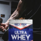 A blurred foreground of a SCI-MX Ultra Whey powder bag with a muscular person holding a dumbbell in the background, set in a gym environment.