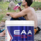 A shirtless man sitting outdoors on a stone path drinks from a pink and clear water bottle. In the foreground, a large blurred container labeled EAA is visible.