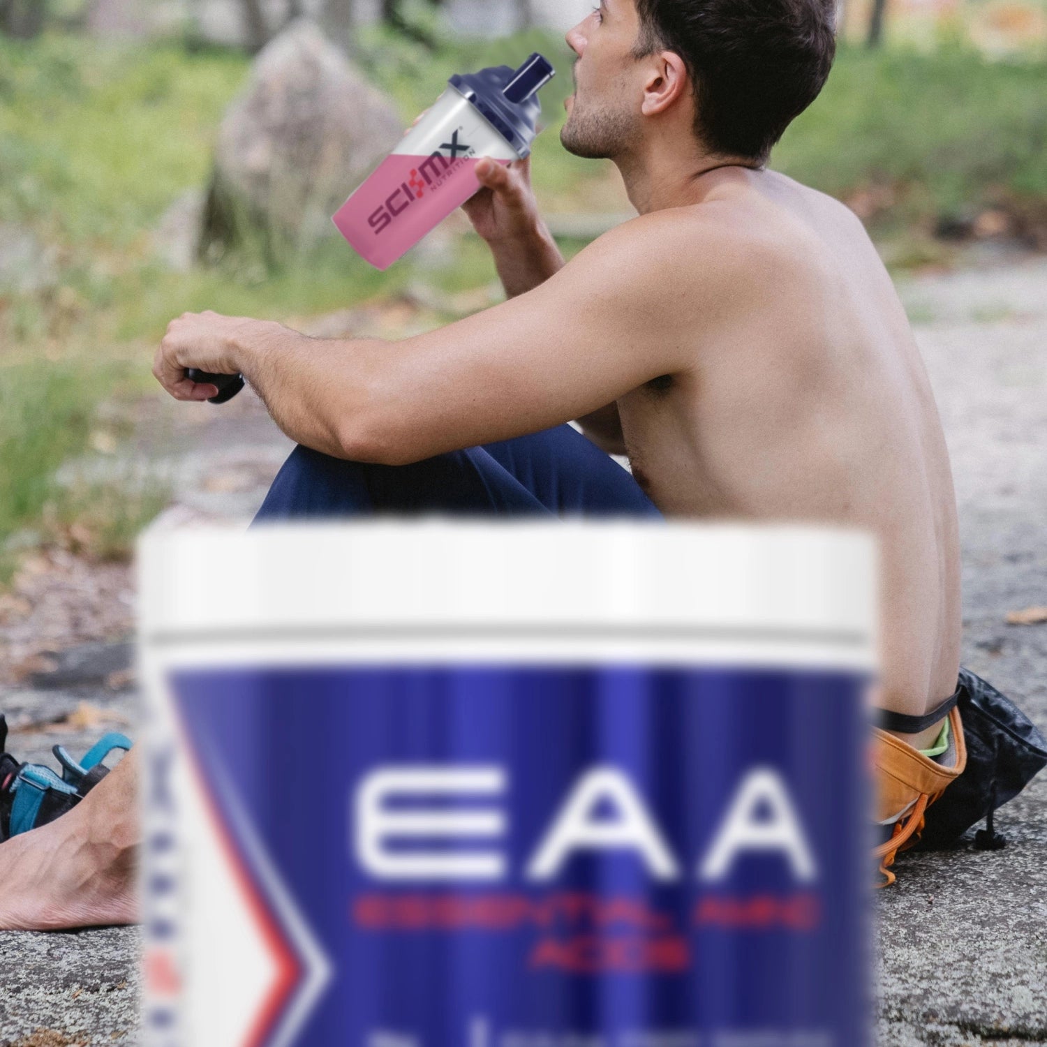 A shirtless man sitting outdoors on a stone path drinks from a pink and clear water bottle. In the foreground, a large blurred container labeled EAA is visible.