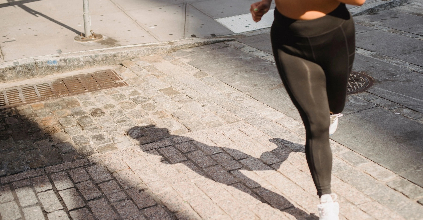 A woman jogging outdoors on a paved sidewalk, wearing black leggings and white sneakers. The image is cropped to show the lower torso and legs in motion, with strong sunlight casting a sharp shadow on the ground.
