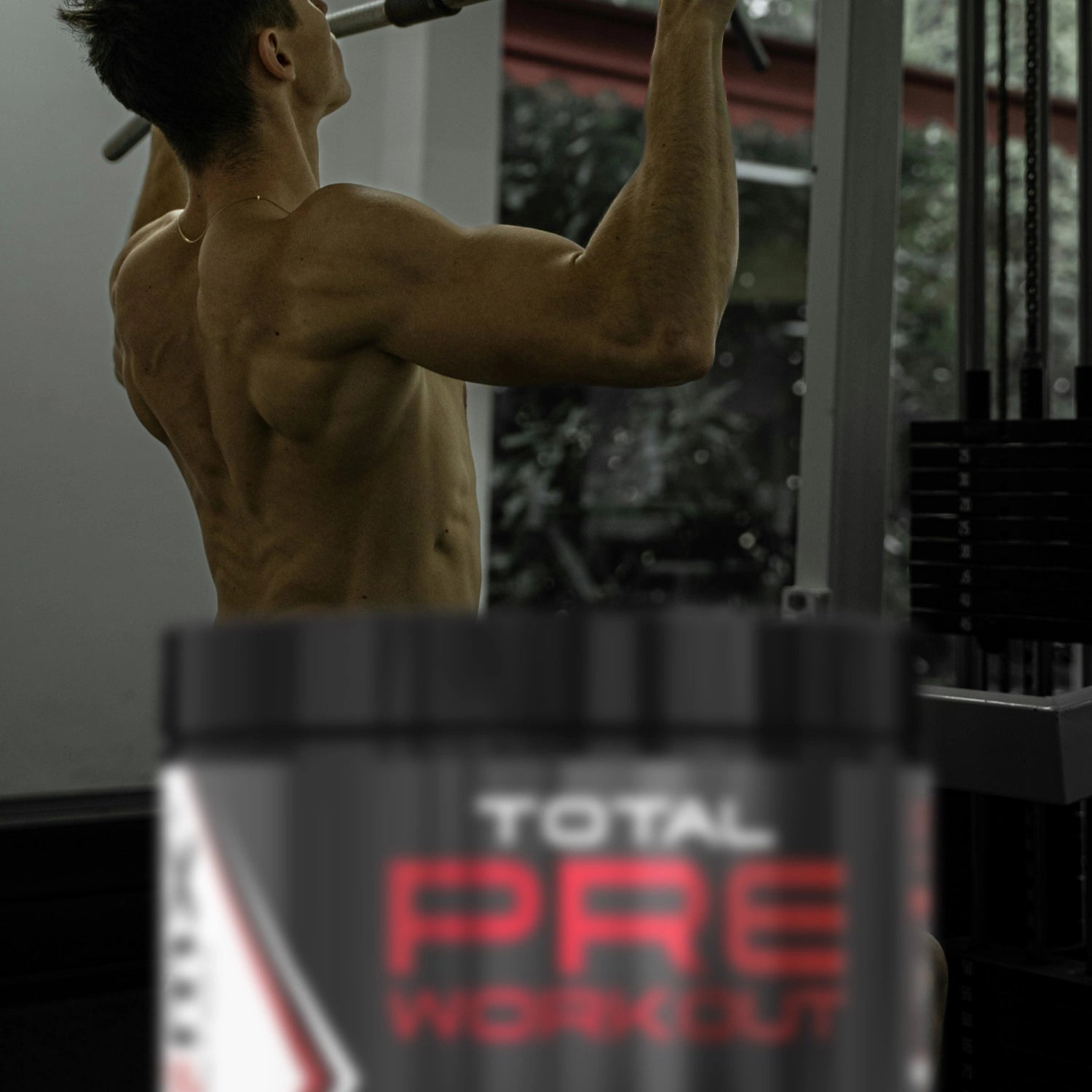 A man doing chin-ups in a gym with a Total Pre Workout supplement container in the foreground.
